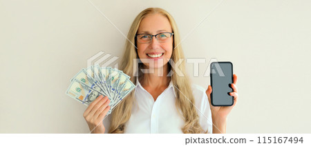 Portrait happy smiling middle-aged woman holding blank screen phone and cash money in dollar bills Portrait happy smiling middle-aged woman holding blank screen phone and cash money in dollar bills 115167494