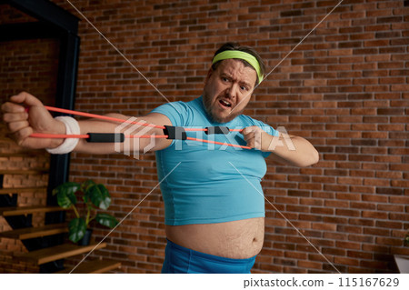 Comic chubby man in sportswear fooling around with fitness equipment Comic chubby man in sportswear fooling around with fitness equipment 115167629