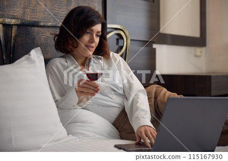Relaxed businesswoman lying in bed drinking wine and browsing internet on laptop Relaxed businesswoman lying in bed drinking wine and browsing internet on laptop 115167930