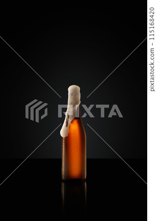 Photo of glass battle filled with amber colored beer, topped with creamy white head against black studio background. 115168420