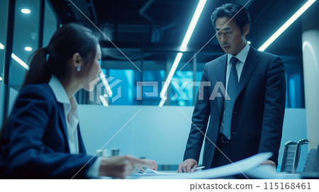 A Japanese businessman is standing at a conference table and looking at a female Japanese colleague explaining a document 115168461