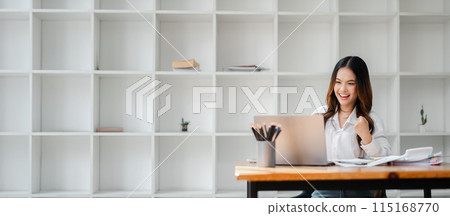 Young woman working on a laptop in a modern office, celebrating success with a smile. Minimalist bookshelf background. Young woman working on a laptop in a modern office, celebrating success with a smile. Minimalist bookshelf background. 115168770