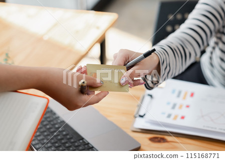 Close-up of hands exchanging a credit card over a wooden table with a laptop and notebook in the background. Close-up of hands exchanging a credit card over a wooden table with a laptop and notebook in the background. 115168771