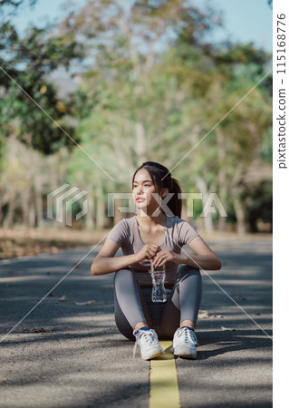 Young woman sitting on a park pathway, holding a water bottle, surrounded by trees and nature, enjoying a sunny day. Young woman sitting on a park pathway, holding a water bottle, surrounded by trees and nature, enjoying a sunny day. 115168776