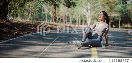 Young woman in athletic clothing resting on a road in a park, with a water bottle, surrounded by trees and nature. 115168777