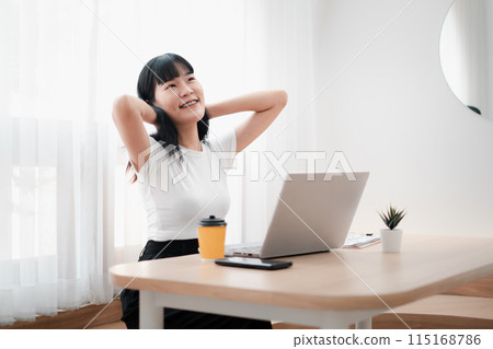 Smiling young woman stretching at her desk with a laptop and coffee, enjoying a break in a bright, modern home office setting. 115168786