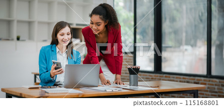Two businesswomen working together in a modern office, using laptops and smartphones, showcasing teamwork and productivity. 115168801