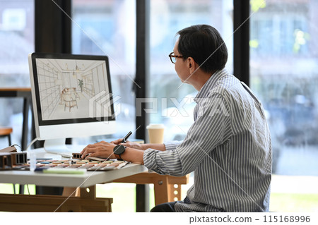 Side view of senior male in eyeglasses working with sketches interior design on computer monitor in office 115168996