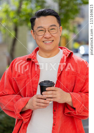 Happy Asian man enjoying drinking morning coffee hot drink, relaxing, taking a break on city street Happy Asian man enjoying drinking morning coffee hot drink, relaxing, taking a break on city street 115169615