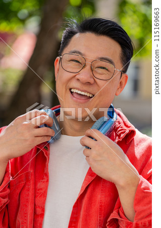 Portrait of happy smiling Asian man listening music taking off headphones looking at camera outdoors 115169663