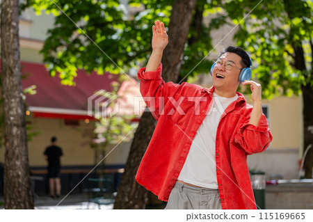 Happy Asian man dancing in wireless headphones listening favorite energetic music on city street Happy Asian man dancing in wireless headphones listening favorite energetic music on city street 115169665
