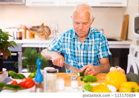 Elderly man cuts vegetables for salad at the table in the kitchen Elderly man cuts vegetables for salad at the table in the kitchen 115170884