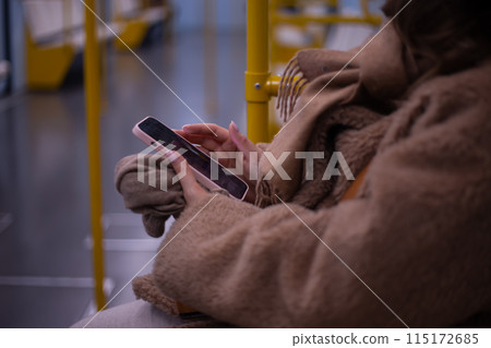 A young woman rides in a modern subway car with a phone in her hands. A beautiful girl in a jacket rides on the subway after work with a smartphone in a post car. Public transport concept 115172685