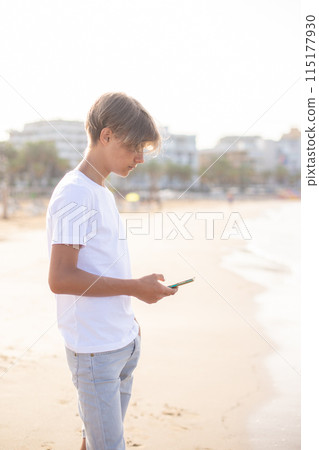 Handsome teenager boy using mobile phone at the beach on summer holidays. Handsome teenager boy using mobile phone at the beach on summer holidays. 115177930