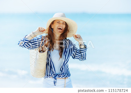 smiling stylish woman on seashore with straw bag 115178724