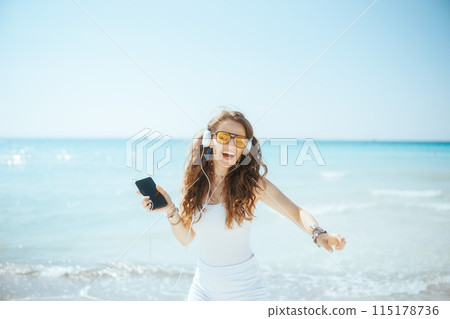 smiling woman on ocean shore dancing and listening to music 115178736