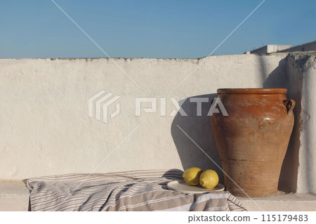 Vintage olive clay storage pot, vase with fresh lemon fruit and textile throw, blanket. White old textured white wall mockup in sunlight. Blue sky background. Summer still life, breakfast composition. 115179483