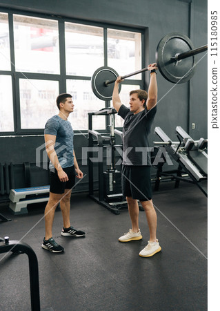 Vertical shot of beginner sportsman exercising at gym support under supervision of coach. Athlete hard time doing barbell overhead press during weightlifting workout with help of personal trainer. Vertical shot of beginner sportsman exercising at gym support under supervision of coach. Athlete hard time doing barbell overhead press during weightlifting workout with help of personal trainer. 115180985