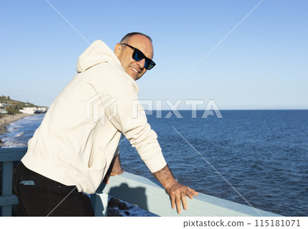 Smiling Middle Age Caucasian Male Standing Alone On Wooden Pier. Blue Sea, Ocean, Sky on Background. Sunny Day. Man Wears Hooded Sweatshirt, Jeans, Sunglasses. Vacation Relaxation. Peaceful Atmosphere Smiling Middle Age Caucasian Male Standing Alone On Wooden Pier. Blue Sea, Ocean, Sky on Background. Sunny Day. Man Wears Hooded Sweatshirt, Jeans, Sunglasses. Vacation Relaxation. Peaceful Atmosphere 115181071