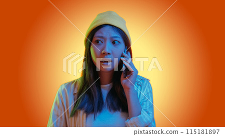 Phone scam. Mobile fraud. Blue color neon light overwhelmed hipster girl talking using gadget isolated on orange gradient copy space background. Phone scam. Mobile fraud. Blue color neon light overwhelmed hipster girl talking using gadget isolated on orange gradient copy space background. 115181897