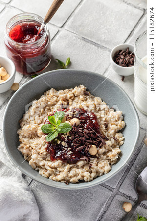 Oatmeal porridge with raspberry jam and nuts in gray bowl on gray tile background served with spoon. Healthy diet breakfast Oatmeal porridge with raspberry jam and nuts in gray bowl on gray tile background served with spoon. Healthy diet breakfast 115182674