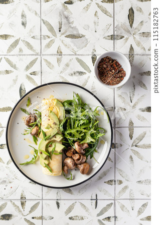 French rolled omelette stuffed with cream cheese with avocado, aragula and mushrooms for breakfast on white plate printed tile background, top view 115182763
