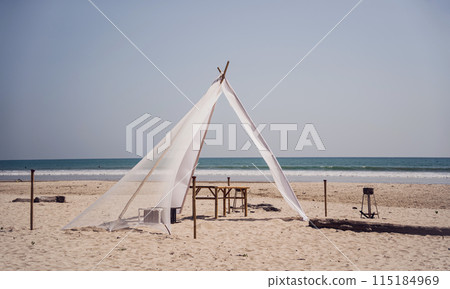 White tent shades sandy beach by ocean under blue sky White tent shades sandy beach by ocean under blue sky 115184969