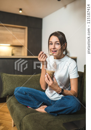 Cute young woman eating dessert from the glass and looking enjoyed 115187414