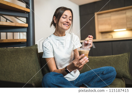 Cute young woman eating dessert from the glass and looking enjoyed 115187415