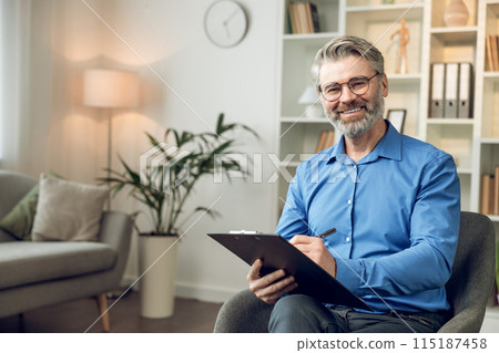 Man psychologist wearing blue shirt holding clipboard standing in his office Man psychologist wearing blue shirt holding clipboard standing in his office 115187458