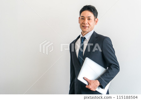 Middle-aged Asian businessman in a suit holding a computer/laptop Middle-aged Asian businessman in a suit holding a computer/laptop 115188504