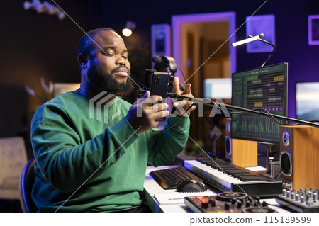 African american songwriter singing his new creation in home studio, recording audio files of his new song for the acoustic version. Young artist musician uses phone to read notes and verses. African american songwriter singing his new creation in home studio, recording audio files of his new song for the acoustic version. Young artist musician uses phone to read notes and verses. 115189599