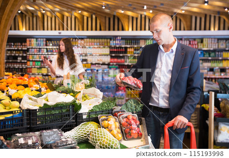 Focused young man in a supermarket selects fresh strawberries 115193998