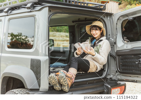 A woman in outdoor fashion reading a book in the luggage compartment of a car while hiking or driving outdoors A woman in outdoor fashion reading a book in the luggage compartment of a car while hiking or driving outdoors 115194322