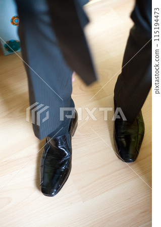 A man in a black suit looks sophisticated while standing on a shiny wooden floor A man in a black suit looks sophisticated while standing on a shiny wooden floor 115194473