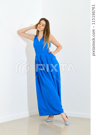 Dreamy woman in long blue sleeveless dress leaning to white wall 115198761