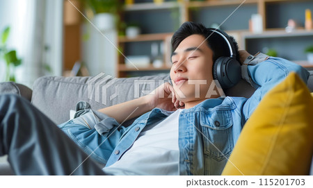 Asian man sitting on couch enjoying to listening to music in wireless headphone, rest and relax. 115201703