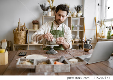 Male artisan sculpting clay in a ceramics workshop with laptop Male artisan sculpting clay in a ceramics workshop with laptop 115202135