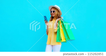 Beautiful happy young woman holding phone with shopping bag, wearing summer hat on blue background 115202851
