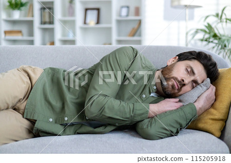 Man taking a nap on a comfortable couch in a well-lit living room. Relaxation and rest in a cozy home environment. 115205918