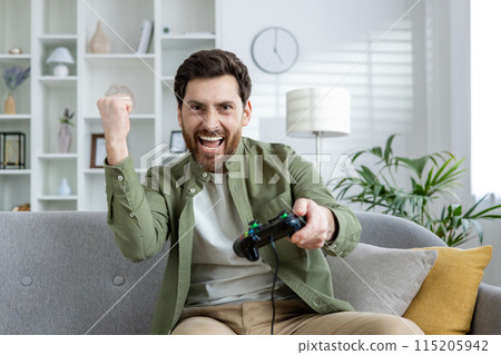 Bearded man enthusiastically playing video games on a couch in a stylish, well-lit living room. He is celebrating a victory while holding a game controller. 115205942