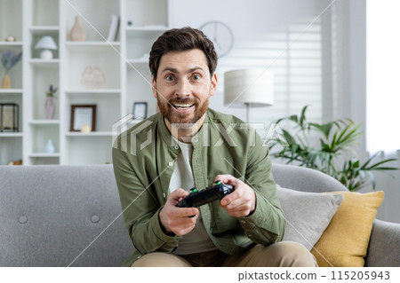 Man playing video game at home, sitting on a comfortable sofa, enjoying leisure time in a modern living room. 115205943