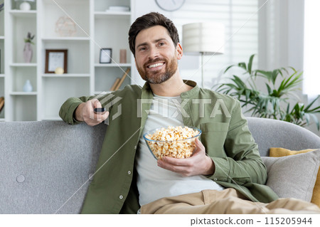 Smiling man sitting on a couch with a bowl of popcorn while watching TV in a cozy living room setting. Smiling man sitting on a couch with a bowl of popcorn while watching TV in a cozy living room setting. 115205944