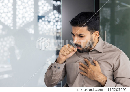 In an office scene, a man with chest discomfort coughs, indicating illness. The detailed background shows frosted glass. Keywords pain, stress, health issues, respiratory problems, concern 115205991
