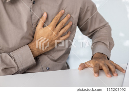 Close-up of a man experiencing chest pain, holding his chest with one hand and resting the other on a table. 115205992