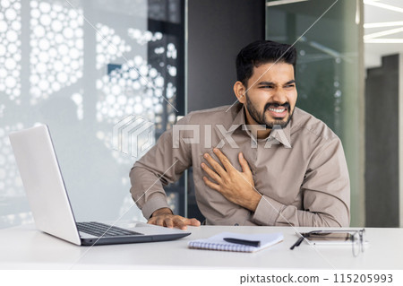 A man sitting at his office desk appears unwell, experiencing chest pain which is causing concern for his health. He is surrounded by gadgets and work materials, trying to cope with his discomfort A man sitting at his office desk appears unwell, experiencing chest pain which is causing concern for his health. He is surrounded by gadgets and work materials, trying to cope with his discomfort 115205993