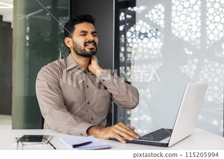 Man in an office setting, sitting at a desk, holding his neck in pain while using a laptop, illustrating discomfort and workplace strain. Man in an office setting, sitting at a desk, holding his neck in pain while using a laptop, illustrating discomfort and workplace strain. 115205994