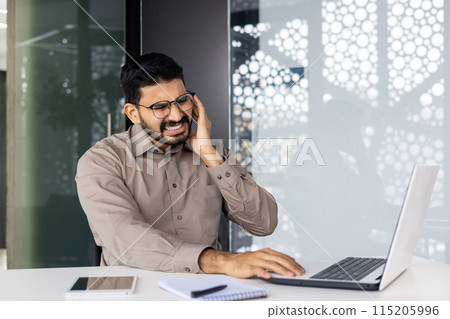 A businessman sitting at his desk, experiencing ear pain and discomfort while working on a laptop. Office environment suggests stress or health issues during work. A businessman sitting at his desk, experiencing ear pain and discomfort while working on a laptop. Office environment suggests stress or health issues during work. 115205996