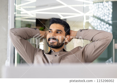 Confident young businessman relaxing with hands behind head while sitting at his desk in a modern office setting, looking satisfied. 115206002