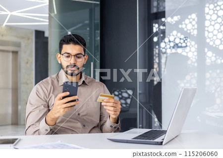 A young businessman in a modern office uses his smartphone and credit card for online shopping. The setting is professional and tech-driven. 115206060